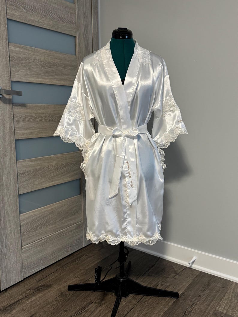 May include: A white satin robe with lace trim. The robe has a long sleeve and a sash tie. The robe is displayed on a mannequin.
