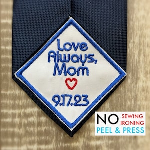 May include: White diamond-shaped iron-on patch with blue stitching. The patch says "Love Always, Mom" with a red heart and the number "917.23".  The text "NO SEWING IRONING PEEL & PRESS" is printed below the patch.