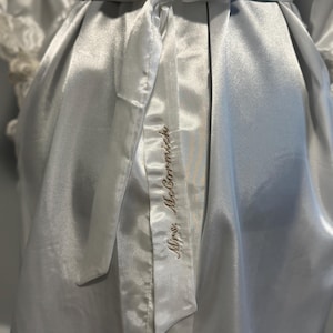 May include: A white satin robe with a white satin sash tied at the waist. The sash has the words 'Mrs. McCormack' embroidered on it in gold thread.
