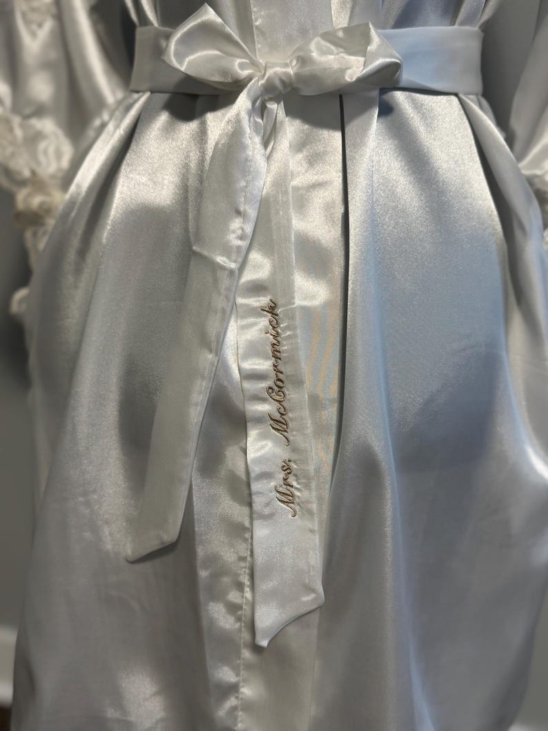May include: A white satin robe with a white satin sash tied at the waist. The sash has the words 'Mrs. McCormack' embroidered on it in gold thread.