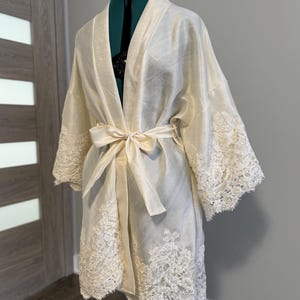 May include: Ivory-colored robe with lace trim on the sleeves and hem, tied with a matching sash. The robe is displayed on a dress form, showcasing its flowing design and delicate details. The lace adds a touch of elegance to this garment.