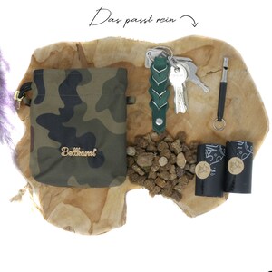 May include: A green camouflage drawstring bag with the word "Bellhamri" in gold lettering, a green braided leather keychain with keys, a whistle, two black dog waste bags, and a pile of brown dog food on a wooden surface.