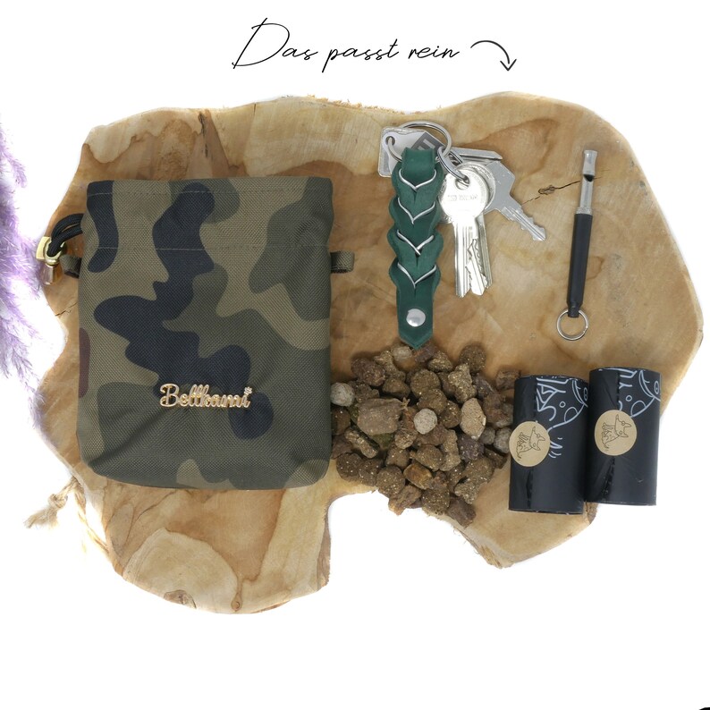 May include: A green camouflage drawstring bag with the word "Bellhamri" in gold lettering, a green braided leather keychain with keys, a whistle, two black dog waste bags, and a pile of brown dog food on a wooden surface.