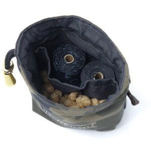 May include: A camouflage-patterned drawstring bag with a gold toggle closure. The bag is open and filled with brown dog treats and two rolls of black dog waste bags.