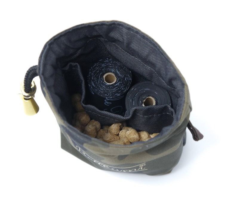 May include: A camouflage-patterned drawstring bag with a gold toggle closure. The bag is open and filled with brown dog treats and two rolls of black dog waste bags.