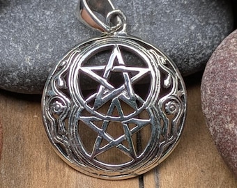 May include: A silver pendant featuring a double pentagram design. The pendant has an ornate, circular frame with decorative swirls and a loop for a chain. The pendant is displayed on a wooden surface with gray and red stones.