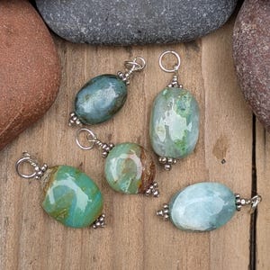 Puede incluir: Cinco colgantes de piedras preciosas pulidas de forma ovalada en tonos verde azulado, verde y azul claro. Cada piedra est&aacute; acentuada con peque&ntilde;as cuentas de metal plateado y una anilla circular en la parte superior, creando piezas de joyer&iacute;a.