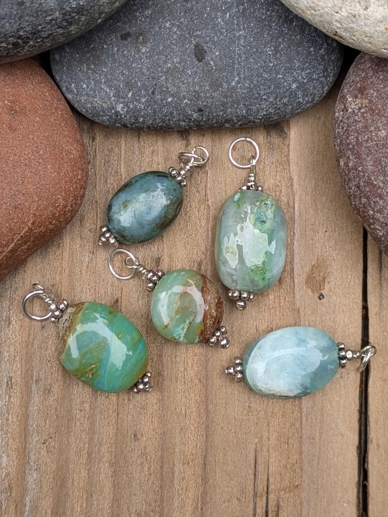 Puede incluir: Cinco colgantes de piedras preciosas pulidas de forma ovalada en tonos verde azulado, verde y azul claro. Cada piedra est&aacute; acentuada con peque&ntilde;as cuentas de metal plateado y una anilla circular en la parte superior, creando piezas de joyer&iacute;a.
