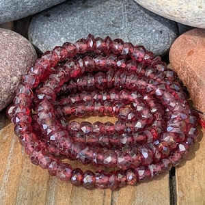 Puede incluir: Un hilo de cuentas de piedras preciosas facetadas de color rojo oscuro, probablemente granate, enrollado sobre una superficie de madera. Las cuentas tienen un aspecto cristalino y brillante, que refleja la luz. El fondo incluye piedras lisas, grises y marrones.
