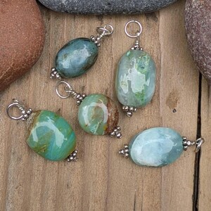 Puede incluir: Cinco colgantes de piedra pulida de forma ovalada en tonos verde azulado, verde y azul claro. Cada colgante cuenta con una anilla de metal plateado en la parte superior y cuentas decorativas de metal plateado en la base, formando un conjunto.