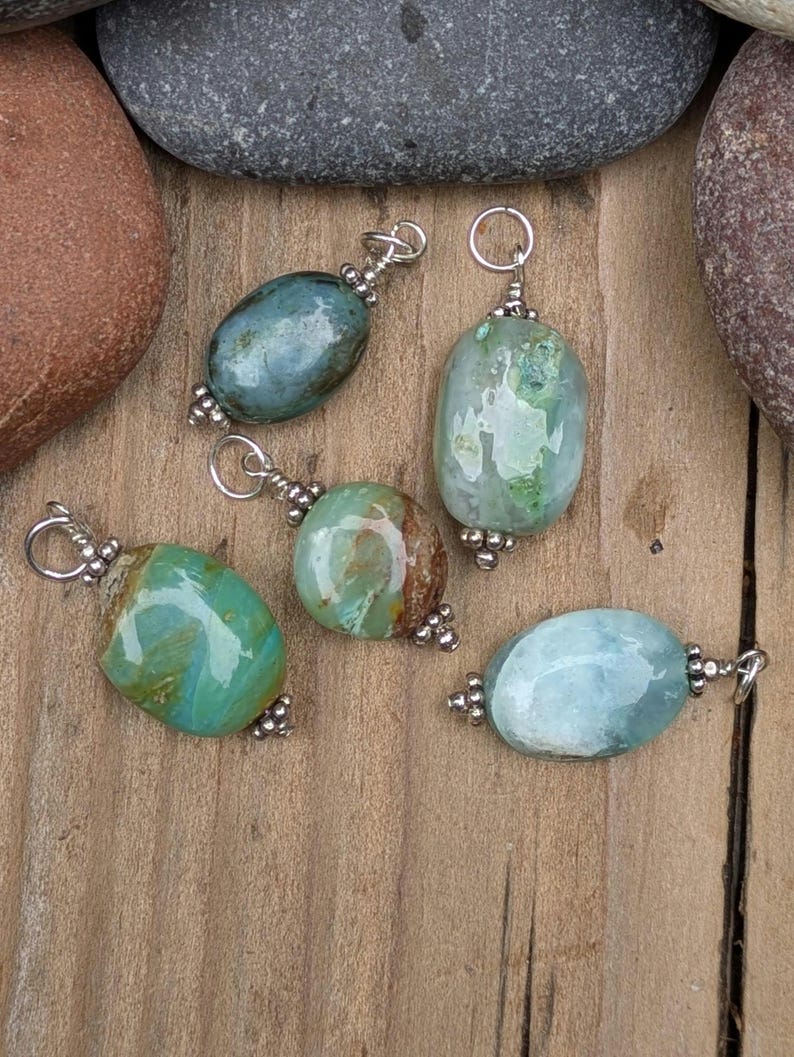 Puede incluir: Cinco colgantes de piedra pulida de forma ovalada en tonos verde azulado, verde y azul claro. Cada colgante cuenta con una anilla de metal plateado en la parte superior y cuentas decorativas de metal plateado en la base, formando un conjunto.