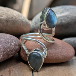 May include: A silver ring with two teardrop-shaped labradorite stones. The ring's band curves around the finger, with one stone at the top and the other at the bottom. The stones display a blue and green iridescence.