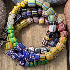 Peut inclure: Un collier de perles de verre colorées et rayées, dans les tons de jaune, bleu, rouge, vert et blanc. Les perles sont enfilées, formant une forme circulaire. Le collier est présenté sur une surface en bois.
