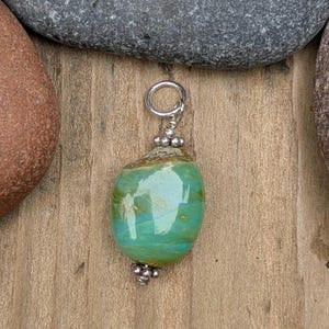Puede incluir: Un colgante con una piedra lisa y pulida de forma ovalada con tonos arremolinados de turquesa, verde y marr&oacute;n. La piedra est&aacute; acentuada con peque&ntilde;as cuentas met&aacute;licas plateadas redondas en la parte superior e inferior, e incluye una anilla de plata.