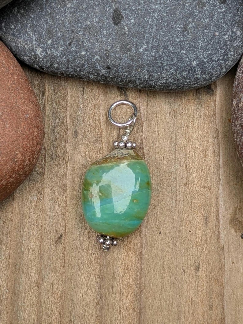 Puede incluir: Un colgante con una piedra lisa y pulida de forma ovalada con tonos arremolinados de turquesa, verde y marr&oacute;n. La piedra est&aacute; acentuada con peque&ntilde;as cuentas met&aacute;licas plateadas redondas en la parte superior e inferior, e incluye una anilla de plata.