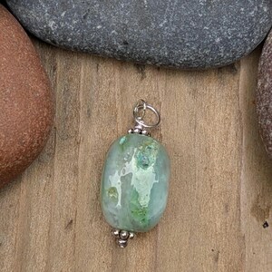 Puede incluir: Un colgante con una piedra ovalada y lisa de color verde claro con variaciones e inclusiones naturales. La piedra est&aacute; decorada con peque&ntilde;as cuentas de metal plateado en la parte superior e inferior, e incluye una argolla de metal plateado.