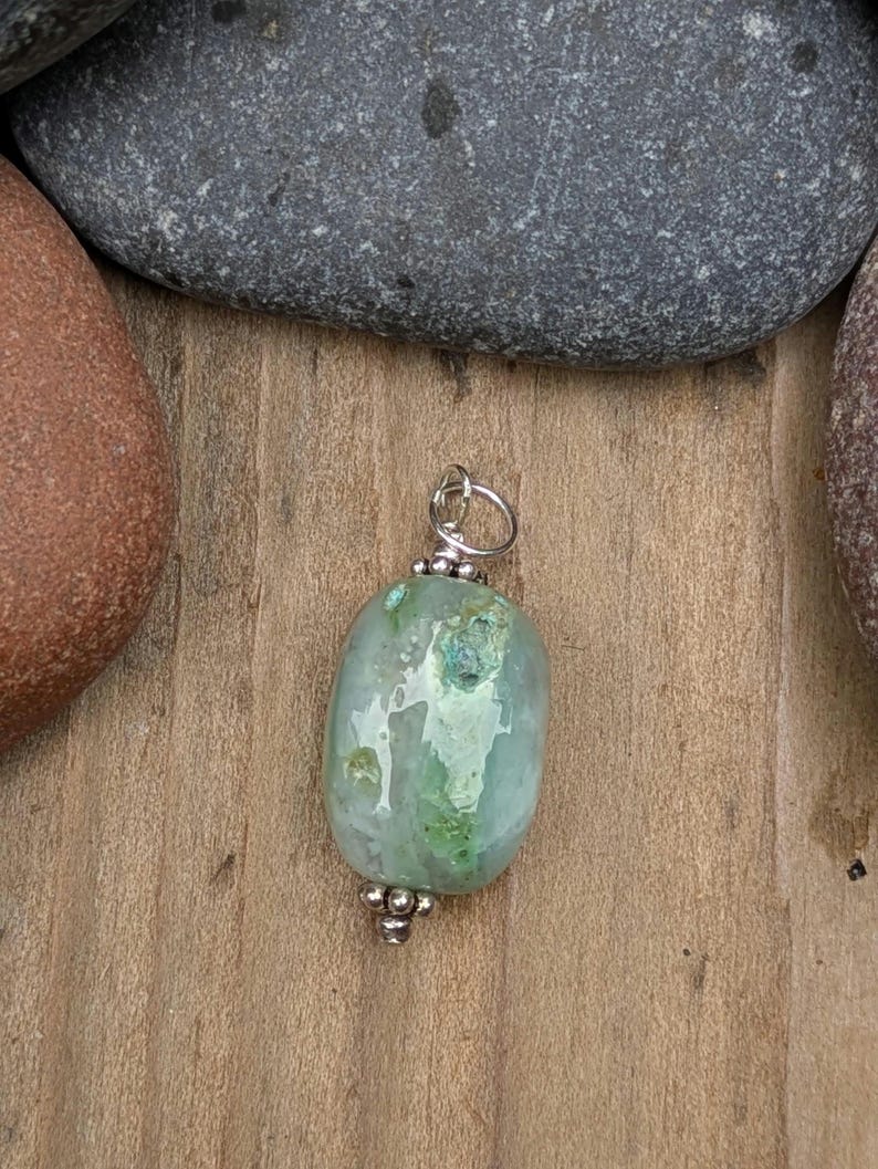Puede incluir: Un colgante con una piedra ovalada y lisa de color verde claro con variaciones e inclusiones naturales. La piedra est&aacute; decorada con peque&ntilde;as cuentas de metal plateado en la parte superior e inferior, e incluye una argolla de metal plateado.