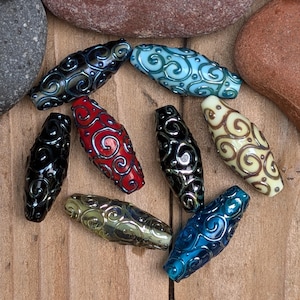 May include: A collection of eight elongated, spindle-shaped beads featuring intricate, raised swirl patterns in metallic silver. The beads come in various colors, including deep blue, light blue, bright red, solid black, cream, and olive green.