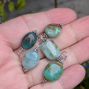 Puede incluir: Cuatro colgantes de piedras preciosas de forma ovalada con superficies pulidas en tonos azul verdoso, azul claro y verde con inclusiones marrones. Cada piedra est&aacute; asegurada con una tapa de metal plateado y una argolla.