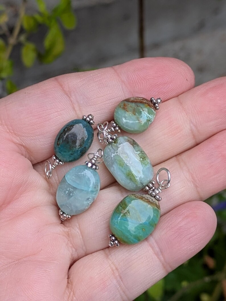 Puede incluir: Cuatro colgantes de piedras preciosas de forma ovalada con superficies pulidas en tonos azul verdoso, azul claro y verde con inclusiones marrones. Cada piedra est&aacute; asegurada con una tapa de metal plateado y una argolla.