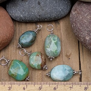 Puede incluir: Cinco colgantes de piedras preciosas pulidas de forma ovalada en tonos verde, azul y marr&oacute;n, cada uno con un enganche de metal plateado con peque&ntilde;as cuentas decorativas. Una regla de madera visible abajo muestra unos 2,5 cm de tama&ntilde;o.