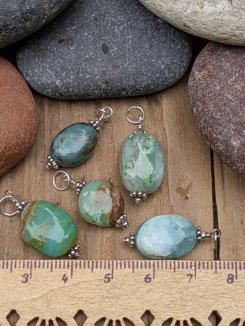 Puede incluir: Cinco colgantes de piedras preciosas pulidas de forma ovalada en tonos verde, azul y marr&oacute;n, cada uno con un enganche de metal plateado con peque&ntilde;as cuentas decorativas. Una regla de madera visible abajo muestra unos 2,5 cm de tama&ntilde;o.