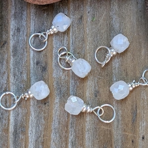 May include: Six small, faceted, clear gemstone charms with silver-colored wire loops and accents. The charms are arranged on a weathered, wooden surface, showcasing their delicate design and potential for jewelry making or crafting projects.