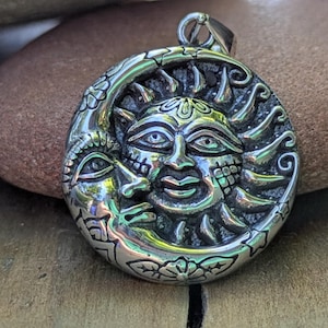 May include: A silver pendant featuring a detailed sun and moon design. The sun has a smiling face with rays, and the moon cradles a smaller face. The pendant is round with floral accents around the edges.