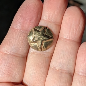 May include: A small, round, gold-colored button with a raised star design in the center. The button has a slightly textured surface and is held in a hand. The star has five points and is the focal point of the button's design.