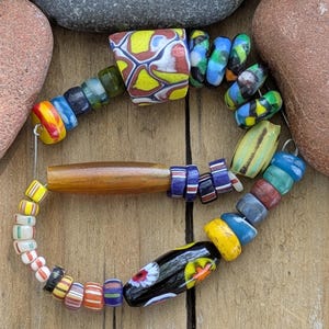 May include: A multi-colored beaded bracelet featuring a variety of glass and wooden beads. The strand includes a large, patterned yellow and red focal bead, a long smooth brown wooden bead, and a black bead with a white and orange floral design.