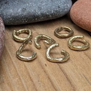 May include: A collection of five small, gold-colored metal ear cuffs with an irregular, textured surface. Each piece features a C-shaped design with a gap, showcasing a hammered or organic finish that reflects light across the metallic surface.