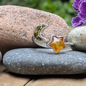 May include: A silver-toned open ring featuring a crescent moon shape with a dark green amber inlay and a five-pointed star shape with a bright orange-yellow amber inlay. The ring band is polished metal and curves to connect the two celestial motifs.