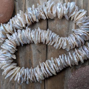 May include: Two strands of iridescent, white keshi pearls are displayed against a rustic wooden background. The pearls have a unique, irregular shape and are strung together with white string. The overall aesthetic is natural and elegant.