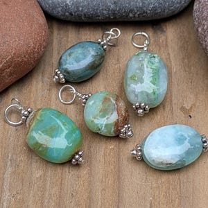 Puede incluir: Cinco colgantes de piedras preciosas de forma ovalada en tonos azul, verde y marr&oacute;n. Cada piedra pulida est&aacute; rematada con cuentas de metal plateado y cuenta con una anilla circular plateada en la parte superior para sujetar a una cadena.