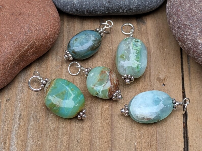 Puede incluir: Cinco colgantes de piedras preciosas de forma ovalada en tonos azul, verde y marr&oacute;n. Cada piedra pulida est&aacute; rematada con cuentas de metal plateado y cuenta con una anilla circular plateada en la parte superior para sujetar a una cadena.