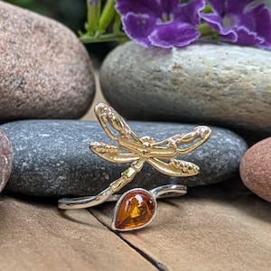 May include: A silver ring featuring a gold-toned dragonfly motif with textured wings and a single teardrop-shaped amber stone set at the base of the band. The design incorporates a bypass style where the dragonfly and the amber stone meet.