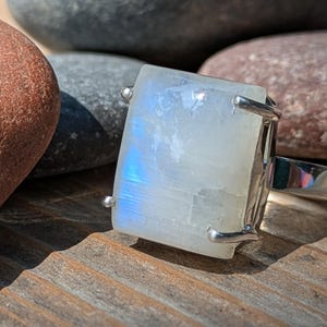 May include: A rectangular moonstone ring with a silver band. The moonstone has a milky white appearance with blue and iridescent flashes. The ring is set on a wooden surface with rocks in the background.