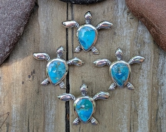 May include: Four silver turtle pendants with turquoise stone inlays. The pendants are arranged on a weathered wooden surface, showcasing the detailed craftsmanship and vibrant blue-green hues of the stones.