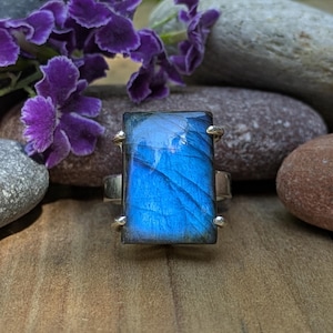 May include: A rectangular labradorite gemstone ring featuring a vibrant blue iridescent flash set in a silver-toned metal band with four prongs holding the stone securely in place, showcasing natural internal fractures and a polished surface.