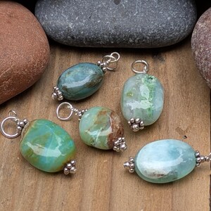 Puede incluir: Cinco colgantes de piedras preciosas de forma ovalada en tonos azul, verde y marr&oacute;n, cada uno con una superficie pulida y un enganche de metal plateado con peque&ntilde;as cuentas decorativas en la parte superior e inferior, sobre una superficie de madera.