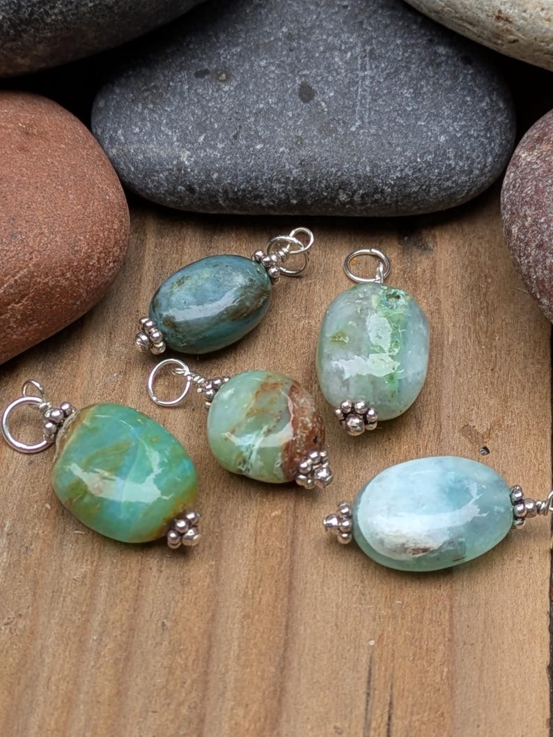 Puede incluir: Cinco colgantes de piedras preciosas de forma ovalada en tonos azul, verde y marr&oacute;n, cada uno con una superficie pulida y un enganche de metal plateado con peque&ntilde;as cuentas decorativas en la parte superior e inferior, sobre una superficie de madera.