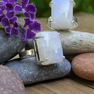 May include: Two silver rings featuring rectangular, translucent moonstone cabochons with a subtle blue adularescence. Each gemstone is secured by four metal prongs on a smooth, polished silver band, showcasing a clean and reflective surface.