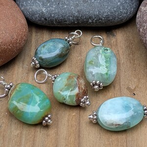 Puede incluir: Cinco colgantes de piedras preciosas pulidas de forma ovalada en tonos verde azulado, verde y azul claro. Cada piedra est&aacute; rematada con cuentas de metal plateado y tiene una peque&ntilde;a anilla plateada en la parte superior para colgar.