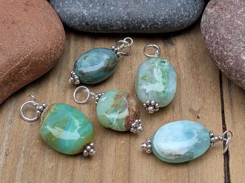 Puede incluir: Cinco colgantes de piedras preciosas pulidas de forma ovalada en tonos verde azulado, verde y azul claro. Cada piedra est&aacute; rematada con cuentas de metal plateado y tiene una peque&ntilde;a anilla plateada en la parte superior para colgar.