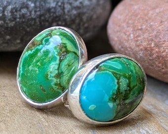 May include: A pair of oval-shaped stud earrings with vibrant green and turquoise stones set in silver. The stones display a marbled pattern with shades of green, blue, and brown. The earrings are displayed on a wooden surface.
