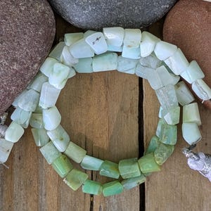 May include: A necklace of light green and white rectangular beads. The beads are strung together, forming a circular shape. The necklace is displayed on a wooden surface, with rocks in the background.