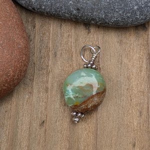 Puede incluir: Un colgante con una piedra pulida, lisa y redondeada, con un patr&oacute;n jaspeado en tonos verde menta, blanco y marr&oacute;n tierra. La piedra est&aacute; sujeta con un alambre plateado, adornado con peque&ntilde;as cuentas met&aacute;licas en la parte superior e inferior.
