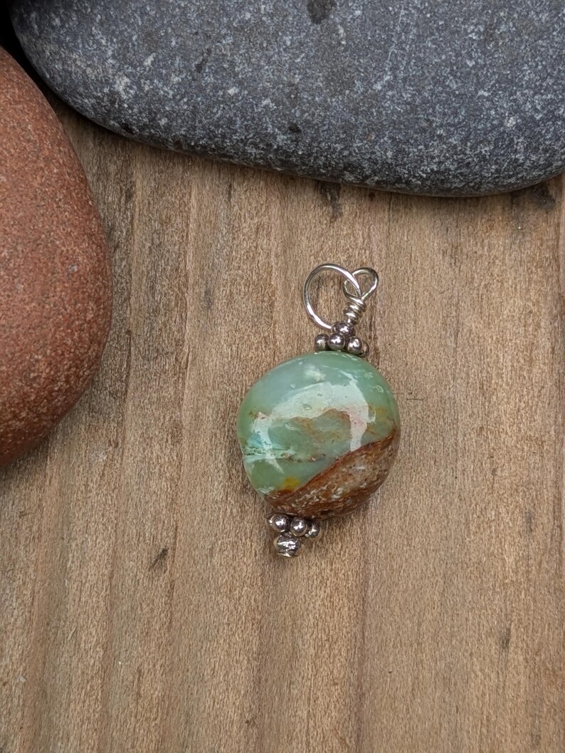 Puede incluir: Un colgante con una piedra pulida, lisa y redondeada, con un patr&oacute;n jaspeado en tonos verde menta, blanco y marr&oacute;n tierra. La piedra est&aacute; sujeta con un alambre plateado, adornado con peque&ntilde;as cuentas met&aacute;licas en la parte superior e inferior.
