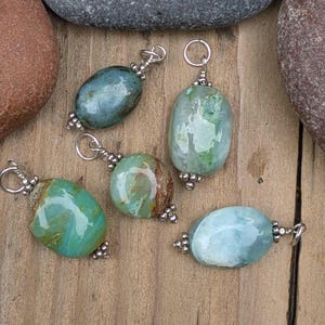 Puede incluir: Cinco colgantes de piedras preciosas pulidas de forma ovalada en tonos azul, verde y marr&oacute;n. Cada piedra est&aacute; envuelta en alambre de tono plateado con una peque&ntilde;a anilla circular en la parte superior y cuentas plateadas decorativas.