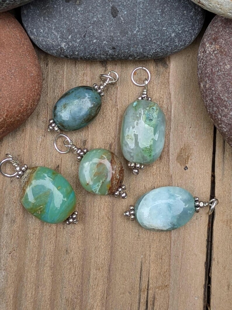 Puede incluir: Cinco colgantes de piedras preciosas pulidas de forma ovalada en tonos azul, verde y marr&oacute;n. Cada piedra est&aacute; envuelta en alambre de tono plateado con una peque&ntilde;a anilla circular en la parte superior y cuentas plateadas decorativas.
